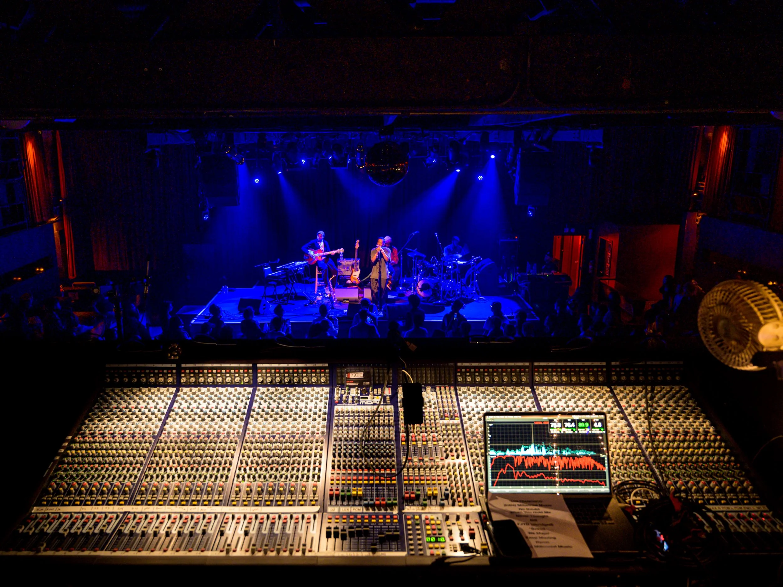 A band on a blue-lit stage seen across a mixing console
